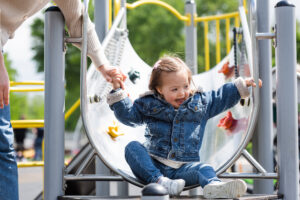 Little girl in a demin jacket and jeans smiling coming to the bottom of a metal slide in a playground holding the hand of an adult woman who is mostly out of shot
