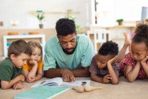A group of small nursery school children with man teacher on floor indoors in classroom, reading books on the floor