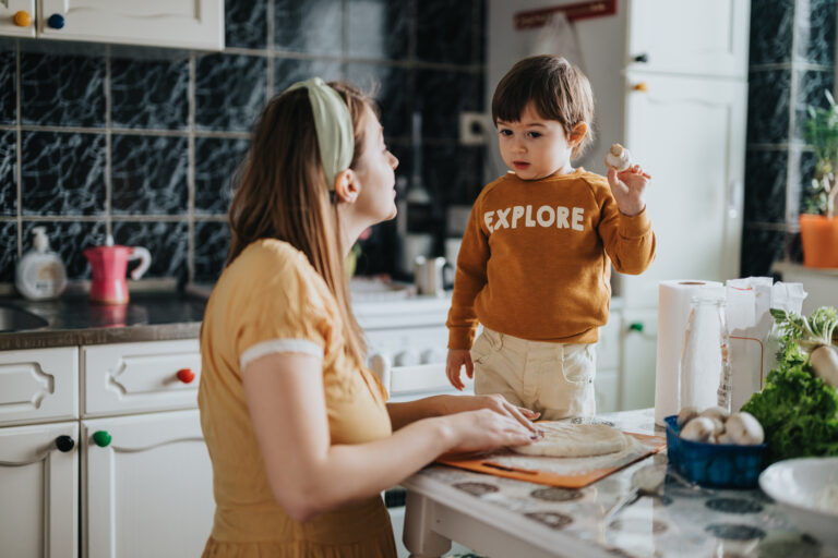Young child in yellow jumper saying 'explore' stands in front of woman in light yellow tshirt kneading dough