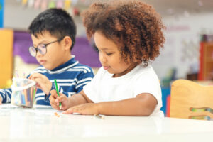 Little girl in a white t-shirt sat a table writing with a green pen next to a little boy wearing glasses and wearing a blue striped t-shirt