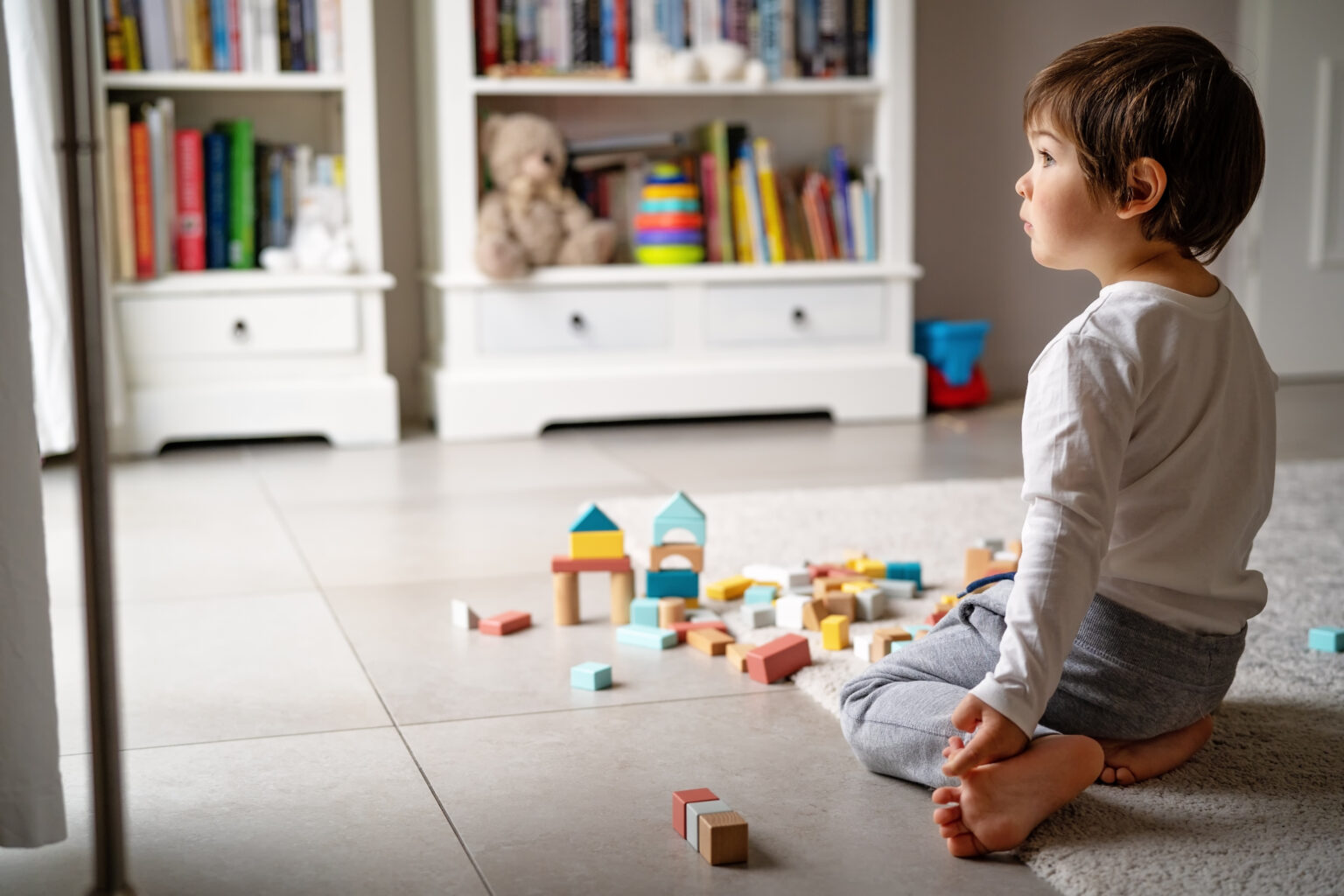 child on tile floor looks int the distance while sat in front of colourful wooden bricks