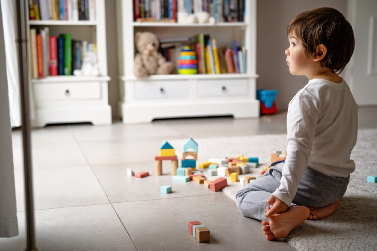 child on tile floor looks int the distance while sat in front of colourful wooden bricks