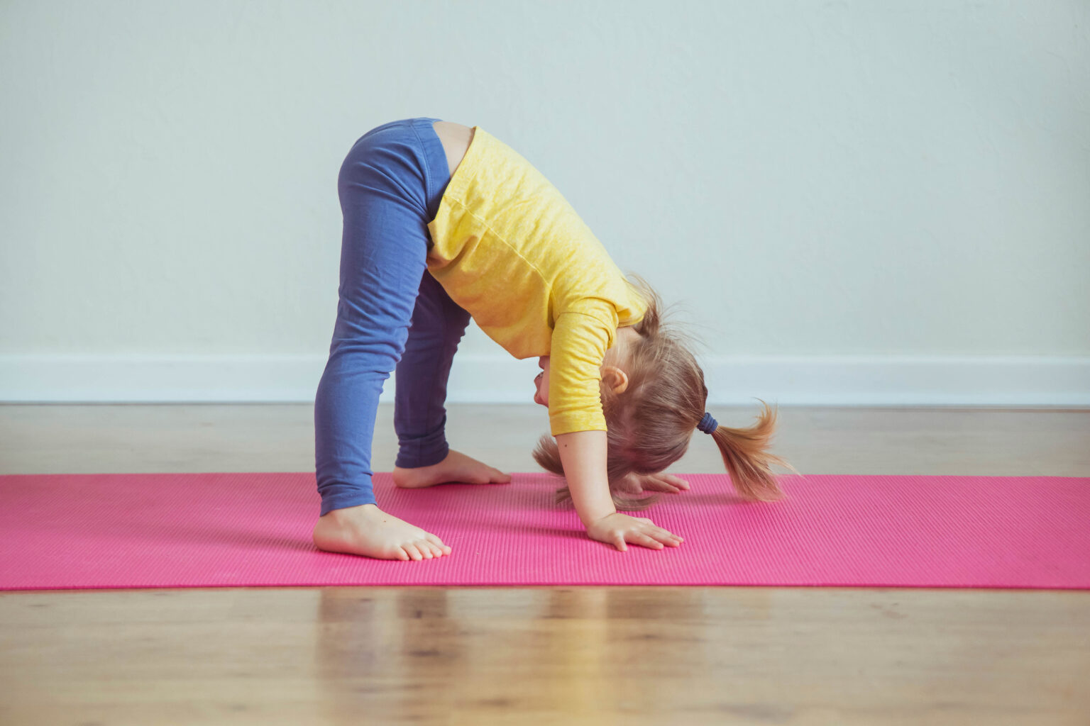 Little girl in a yellow t-shirt and jeans doing a forward yoga pose on a pink mat